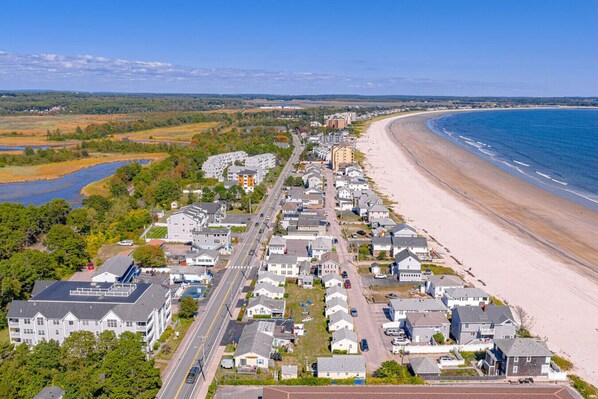 A coastal residential area with houses along a beach, a road, and a river in the background.