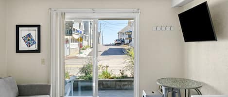 A compact living space with a sofa, a small table, a kitchenette, and a sliding glass door leading to a street view.