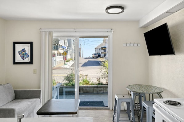 A compact living space with a sofa, a small table, a kitchenette, and a sliding glass door leading to a street view.
