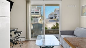 A compact living space with a sliding glass door leading to a balcony, a small dining table, a sofa, and a kitchen area with a refrigerator.