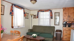 Uma sala de estar acolhedora com um sofá verde, mesa de jantar de madeira e cadeiras. Há uma pequena área de cozinha com armários de madeira e uma lixeira.