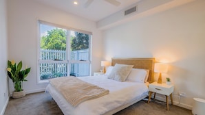 A bedroom with a large bed, two bedside tables, a window with a view of a white fence and greenery, and a ceiling fan. - Cedar Sands Hideaway - 3BRM Home (Gold Coast)