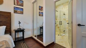 Une chambre à coucher comprenant un lit, une table de chevet, un bureau et un grand miroir. Une porte-fenêtre coulissante donne accès à une salle de bain dotée d’une cabine de douche en verre.