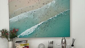Zona pranzo con tavolo in legno, sottotavolo intrecciato e un'ampia opera d'arte incorniciata raffigurante una spiaggia.