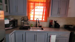 A kitchen with grey cabinets, a microwave, a sink, and a window with orange curtains. - Silverbuttons  (St. John's)