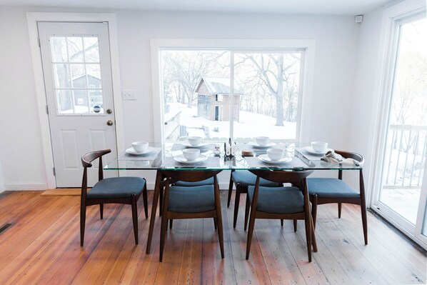 A dining room with a glass table and six chairs, a white door, and a view of a house through large windows.