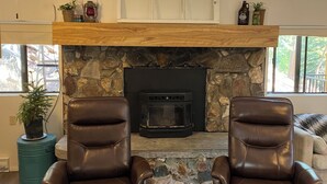 Una sala de estar con chimenea de piedra, dos sillones de cuero, un repisa de madera y un ventilador de techo.