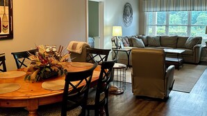 Uma sala de jantar com mesa de madeira, cadeiras e um candelabro. Há um sofá e um abajur na sala de estar.