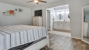 Un dormitorio con cama, ventilador de techo, televisión, cómoda y baño con tocador y espejo.