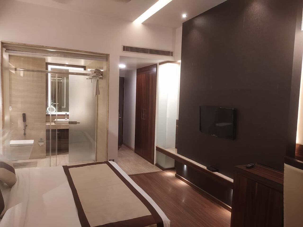 A modern hotel room with a glass shower enclosure, a flat-screen TV mounted on the wall, and a wooden floor.