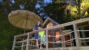 A wooden deck with a rope railing, a thatched umbrella, and a rustic building with a lit interior. - Charming Waterfront Tree House in Kaufman (Kaufman)