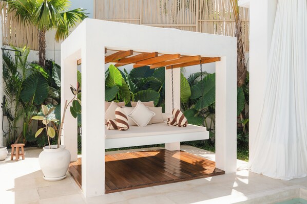 A white outdoor seating area with a wooden swing bed, surrounded by tropical plants.