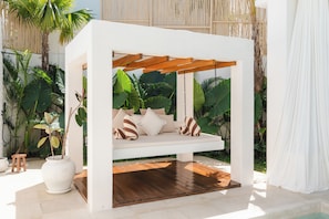 A white outdoor seating area with a wooden swing bed, surrounded by tropical plants.