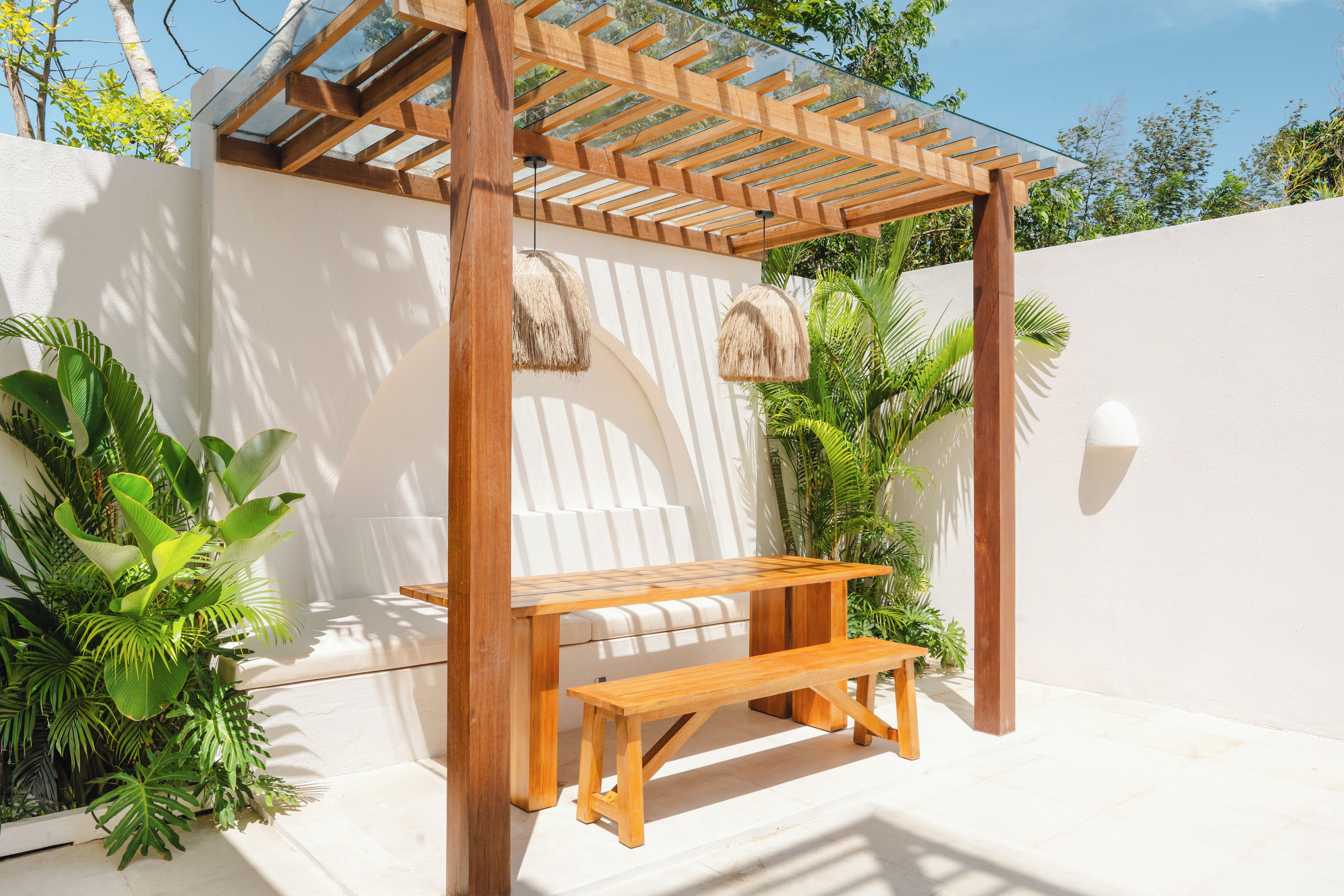 A wooden pergola with a bench, surrounded by lush green plants and a white wall.