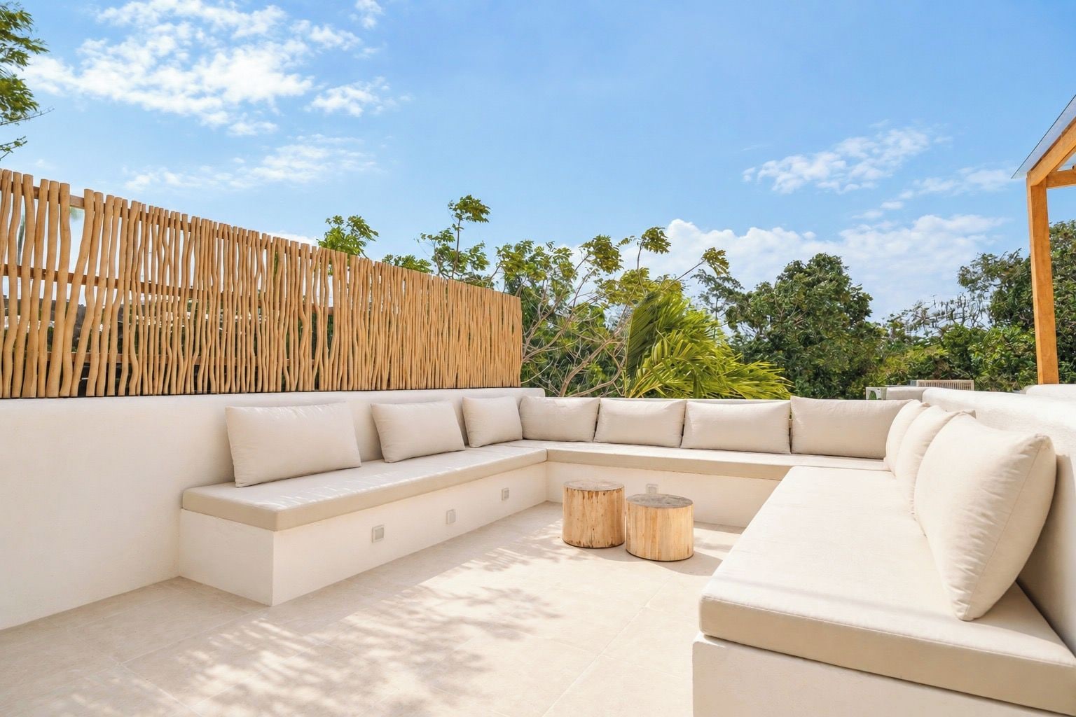 An outdoor seating area with a curved sofa and round wooden tables, surrounded by a wooden fence and lush greenery.
