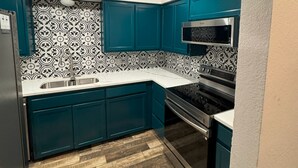 A compact kitchen with teal cabinets, a stainless steel microwave, and a wood-patterned backsplash.