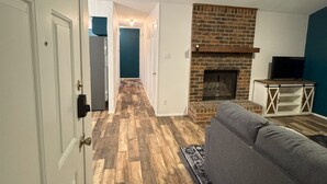 A living room with a brick fireplace, a grey sofa, and a wooden floor.