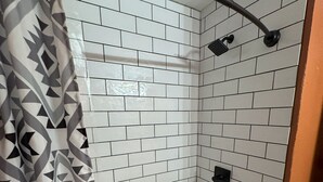 A bathroom with white tiled walls, a shower with a black handle, and a white bathtub.