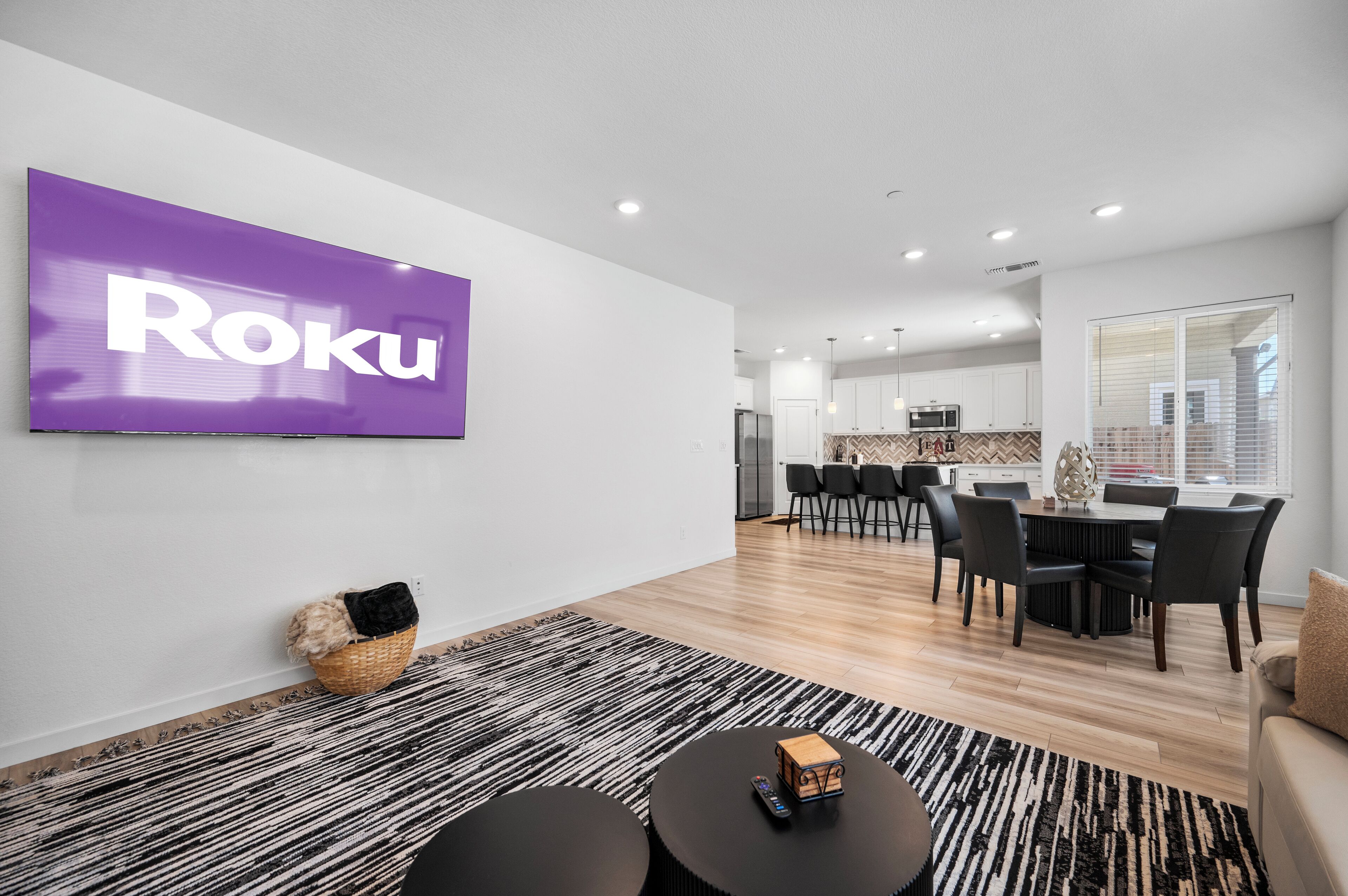 Ein modernes Wohnzimmer mit einem großen Flachbildfernseher, der "Roku" anzeigt, ein Essbereich mit einem schwarzen Tisch und Stühlen sowie eine Küche im Hintergrund.