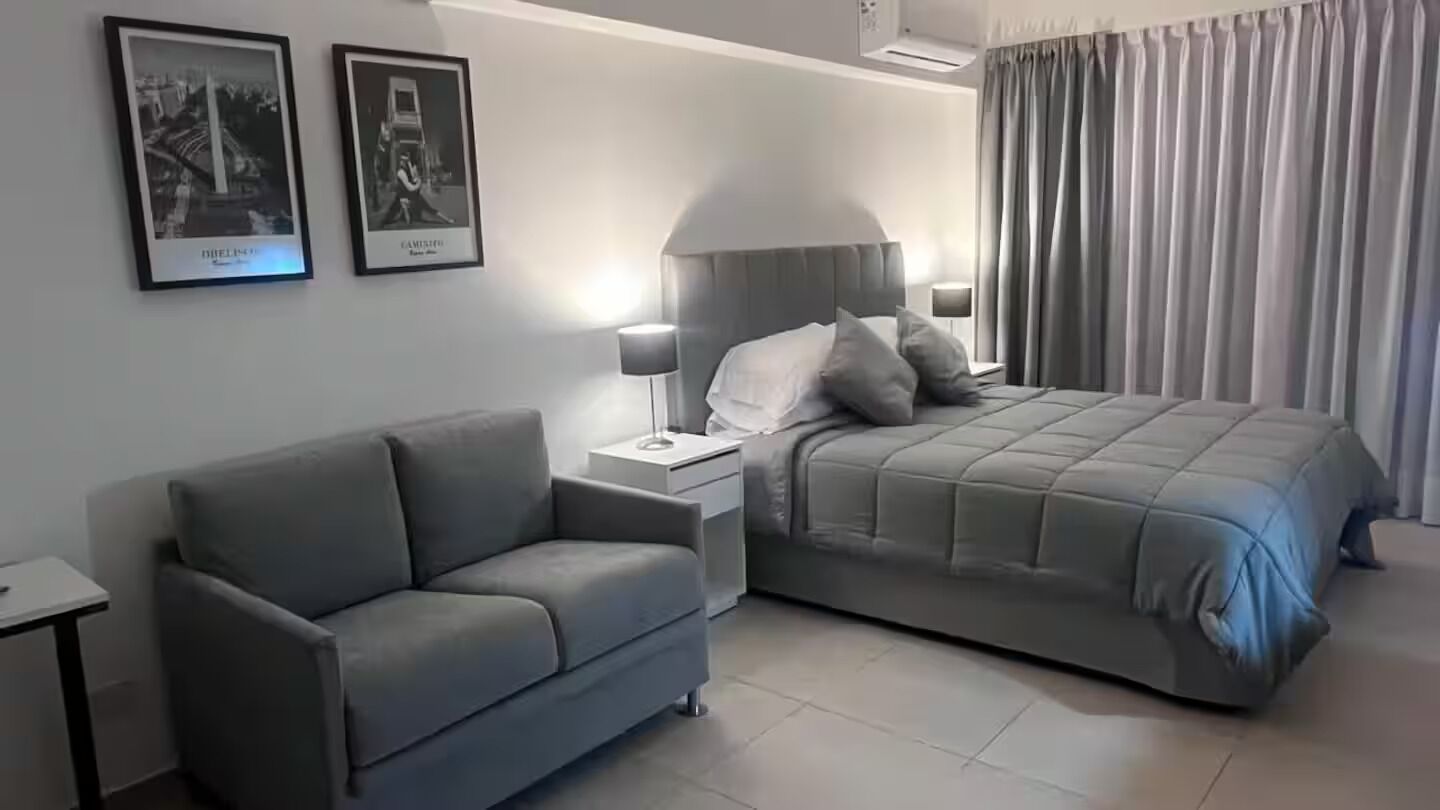 A modern hotel room with a grey sofa, a bed with a grey duvet, and two framed pictures on the wall.