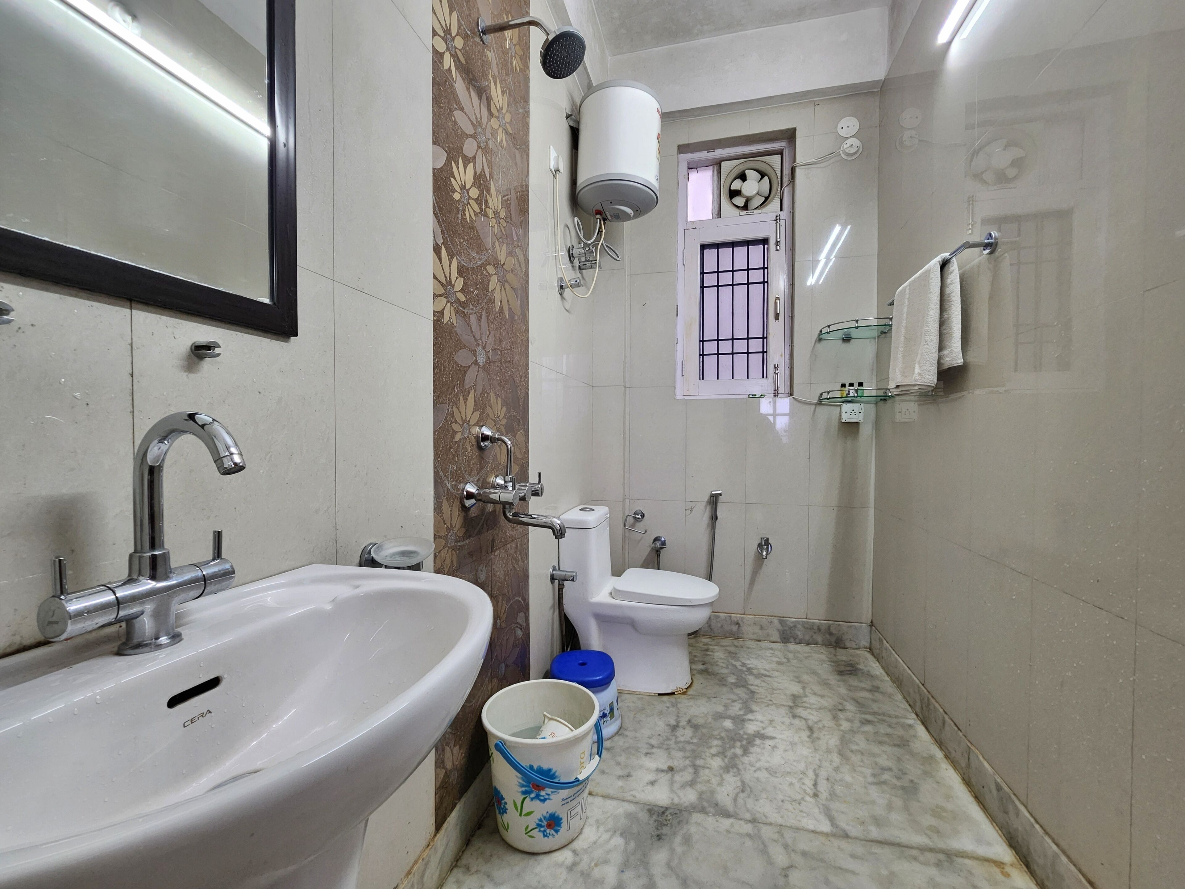 Une salle de bain équipée d’un lavabo, de toilettes et d’une douche. On y trouve une fenêtre avec des barreaux, un ventilateur et un porte-serviettes.