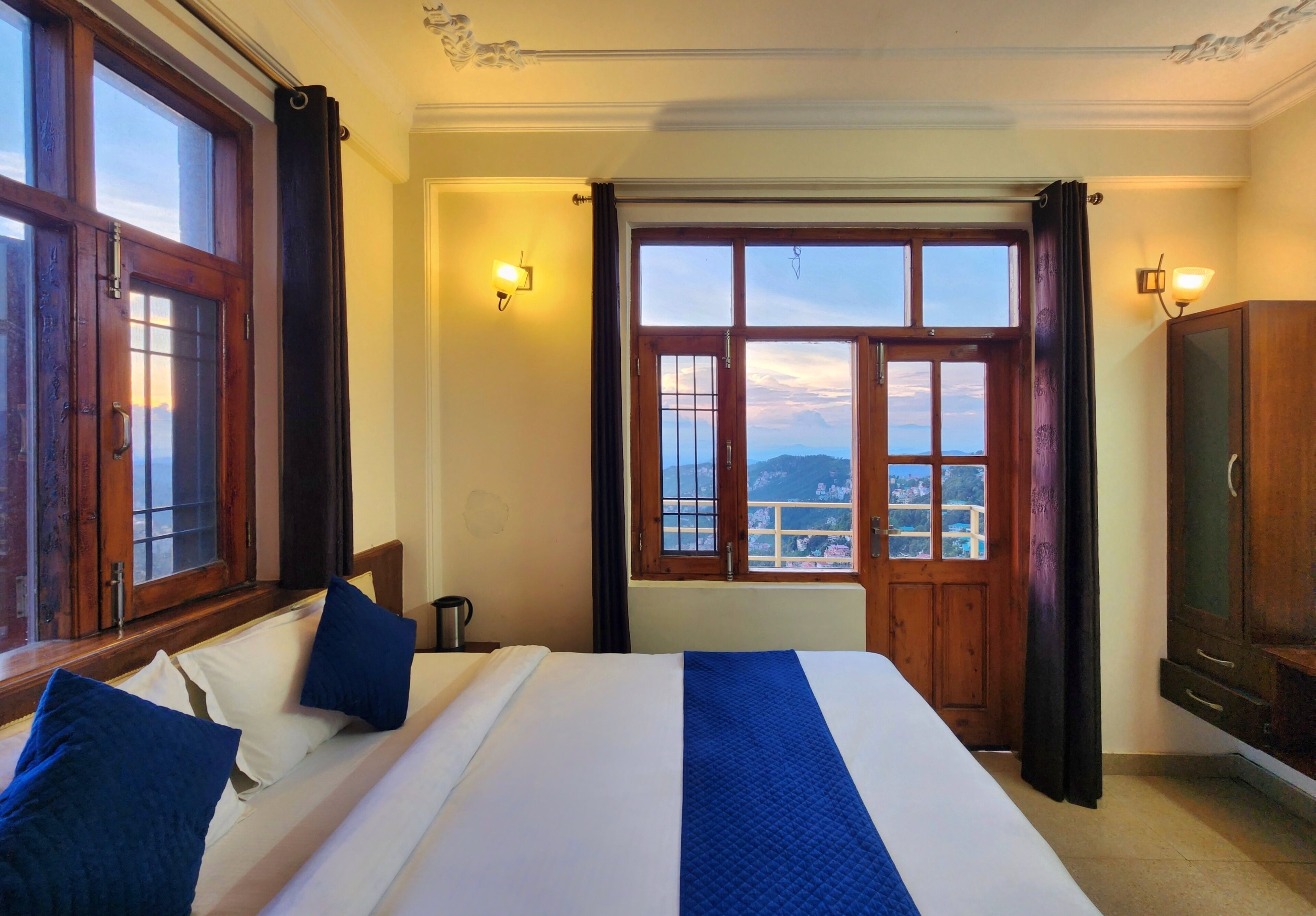 A hotel room with a large bed, a view of the city through a window, and a wooden wardrobe.