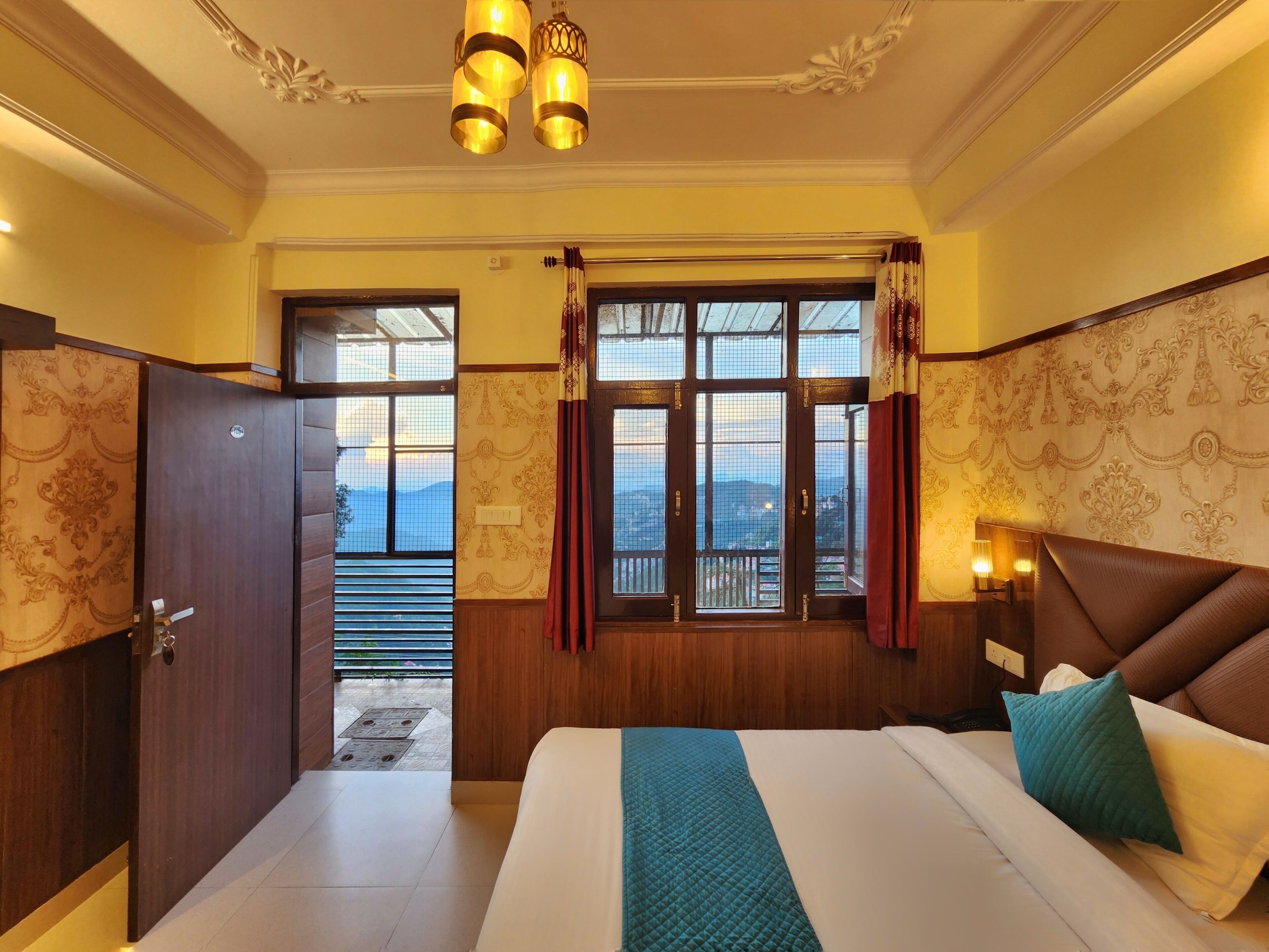 A hotel room with a bed, a nightstand, a window with curtains, a door, and a view of the outdoors.