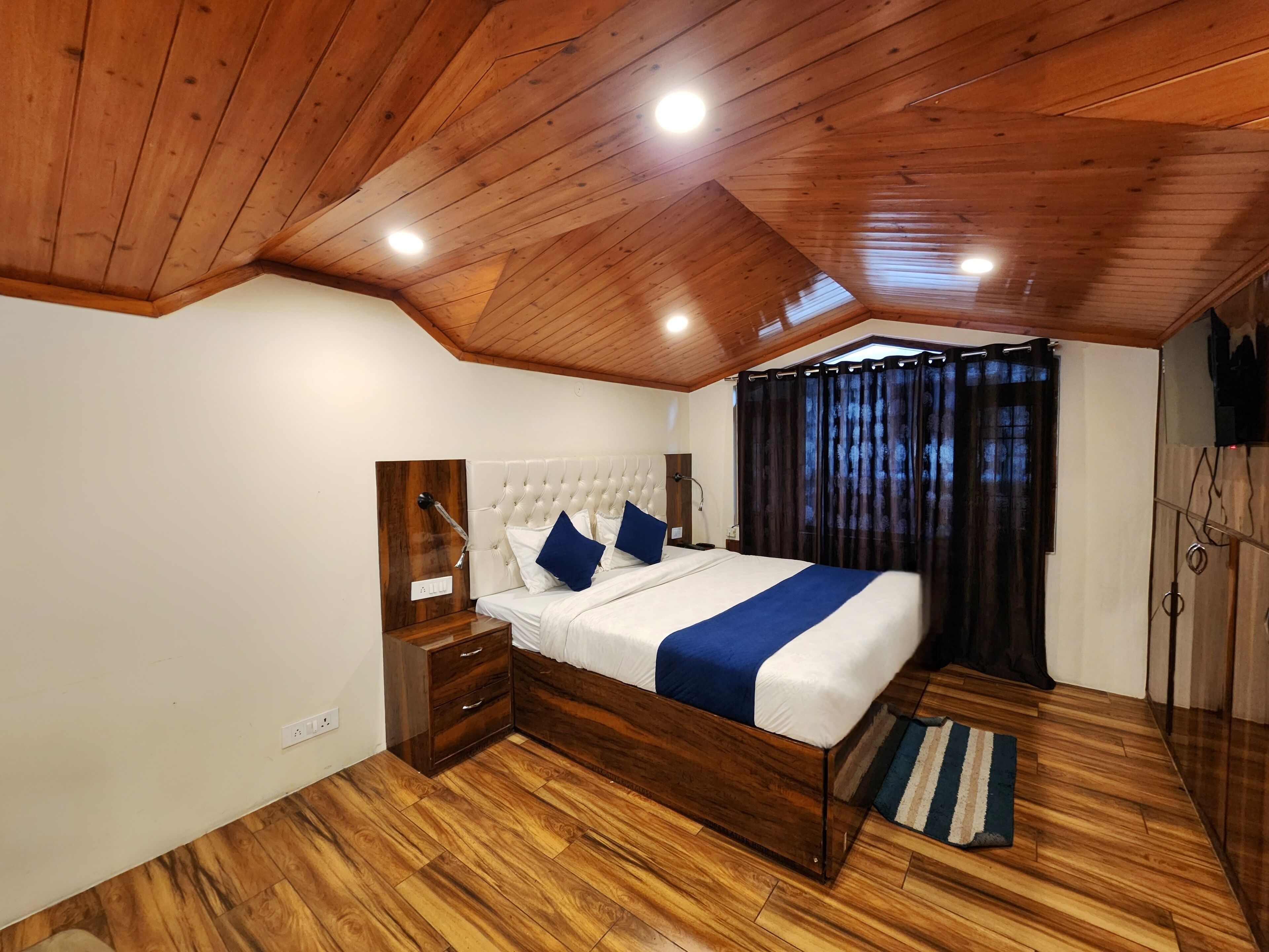 A bedroom with a wooden ceiling, a bed with white and blue bedding, a nightstand, a wooden dresser, and a window with curtains.