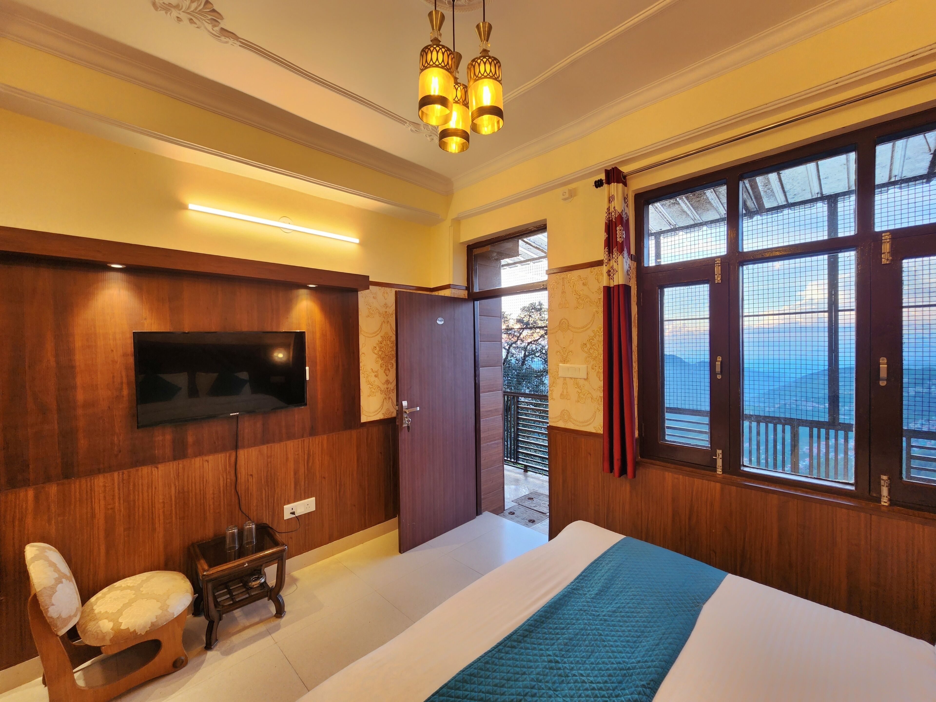 A hotel room with a wooden panel wall, a flat-screen TV, a bed with a blue quilt, a wooden chair, and a balcony with a view.