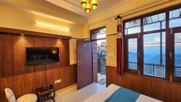 A hotel room with a wooden panel wall, a flat-screen TV, a bed with a blue quilt, a wooden chair, and a balcony with a view.