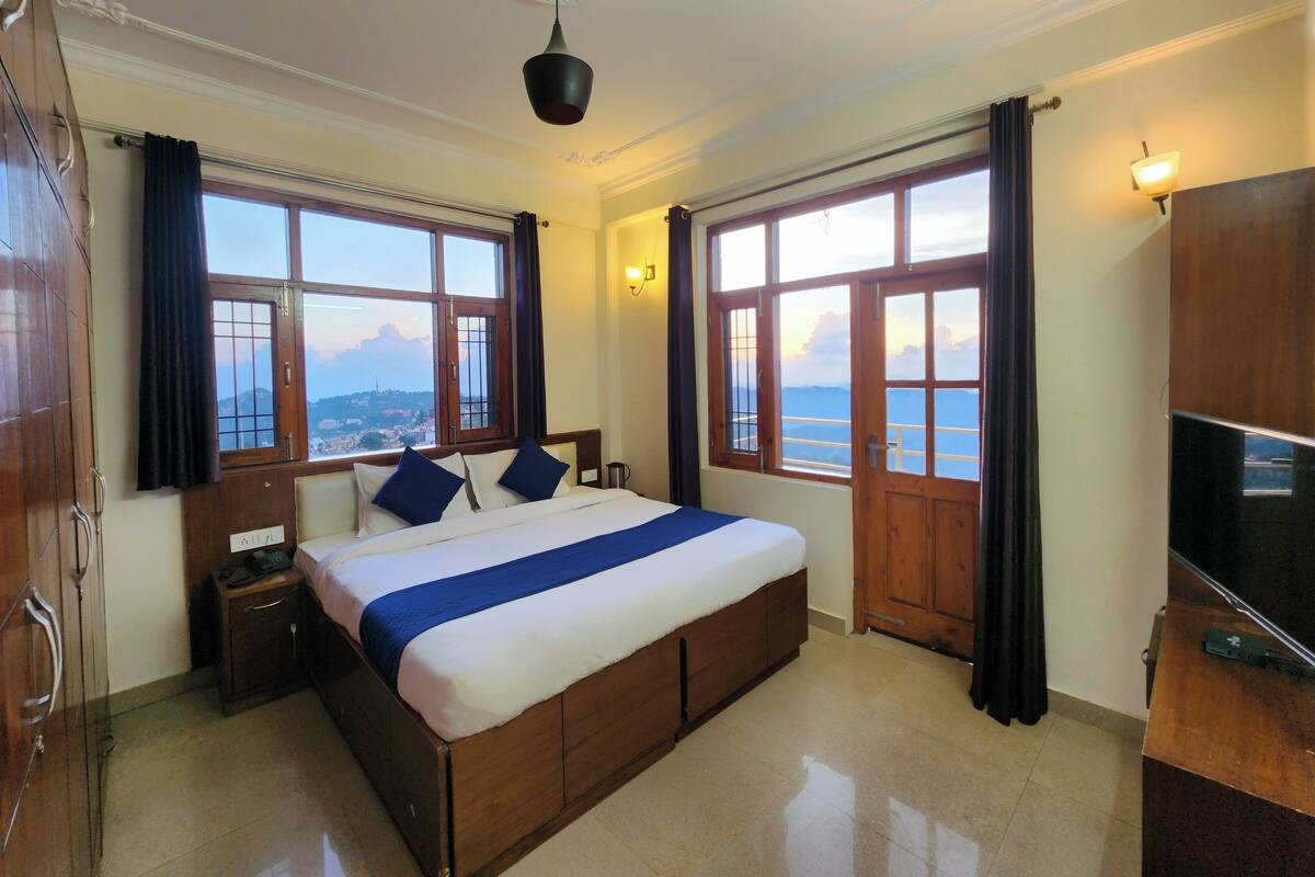 A bedroom with a large bed, wooden headboard, two bedside tables, a nightstand, a TV, and a view of the sea through large windows.