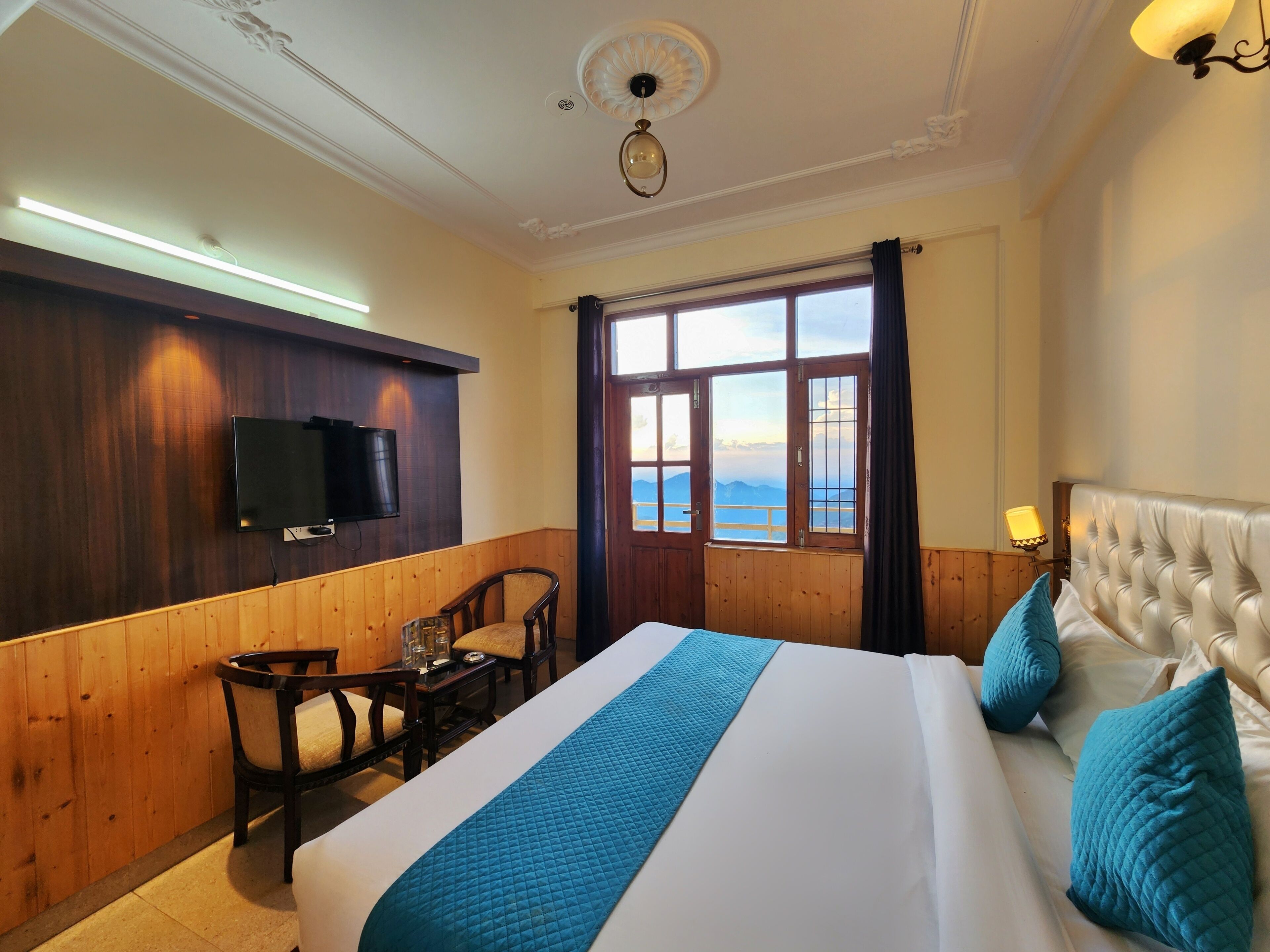 Habitación de hotel con una cama grande, televisión instalada en la pared, una mesa de madera con sillas y una ventana con vistas a las montañas.