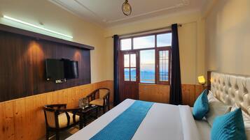 Habitación de hotel con una cama grande, televisión instalada en la pared, una mesa de madera con sillas y una ventana con vistas a las montañas.
