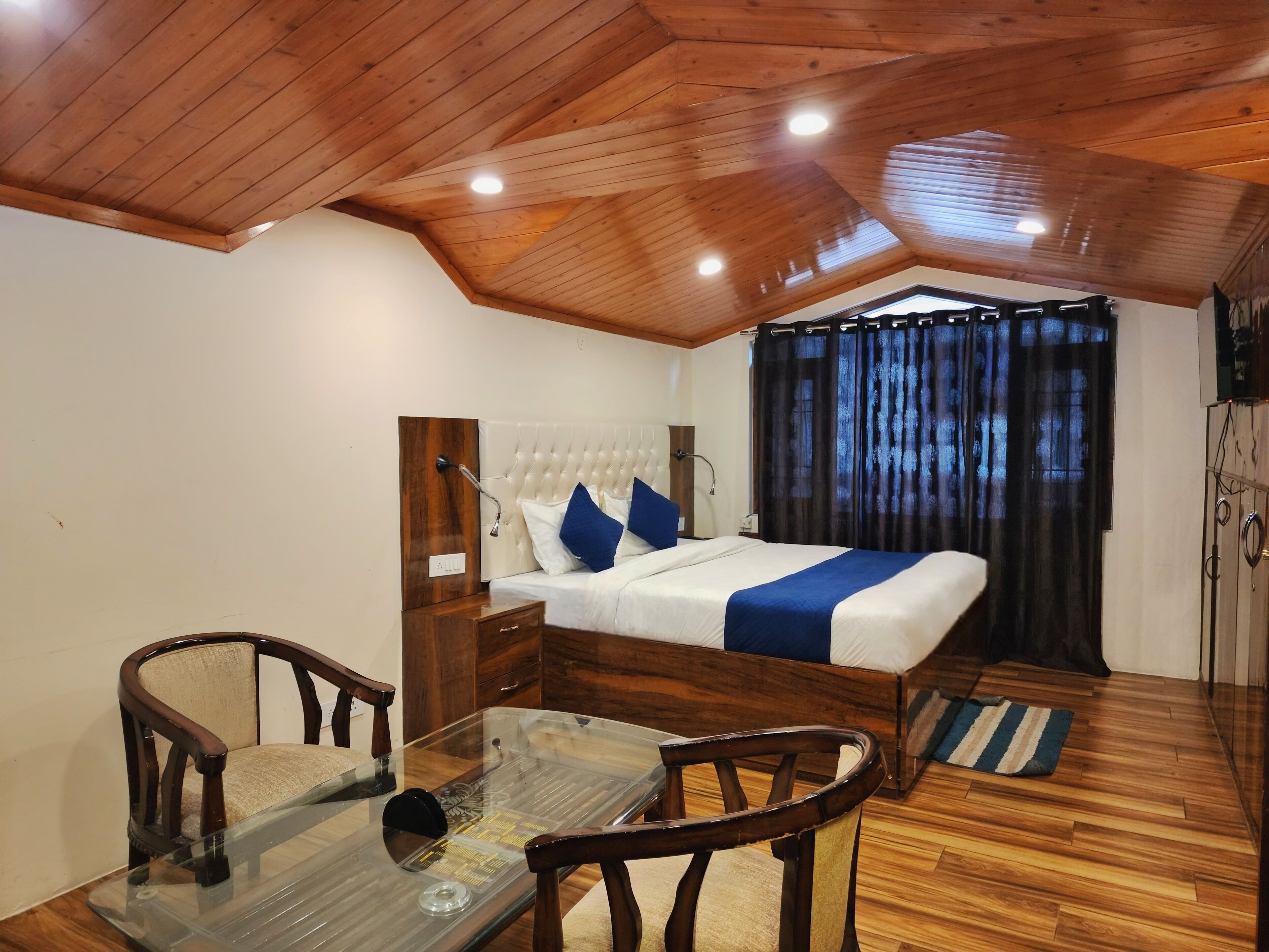 A bedroom with a wooden ceiling, a bed with white bedding and blue pillows, a glass table with chairs, and a wooden floor.