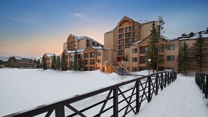 Un espace extérieur enneigé, délimité par une clôture en bois, avec un bâtiment à étages et doté de balcons, et un ciel dégagé.