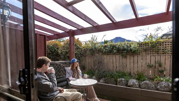 Un espace extérieur abrité, aménagé avec une table et des chaises, une terrasse en bois et offrant une vue sur un jardin et des montagnes en arrière-plan.