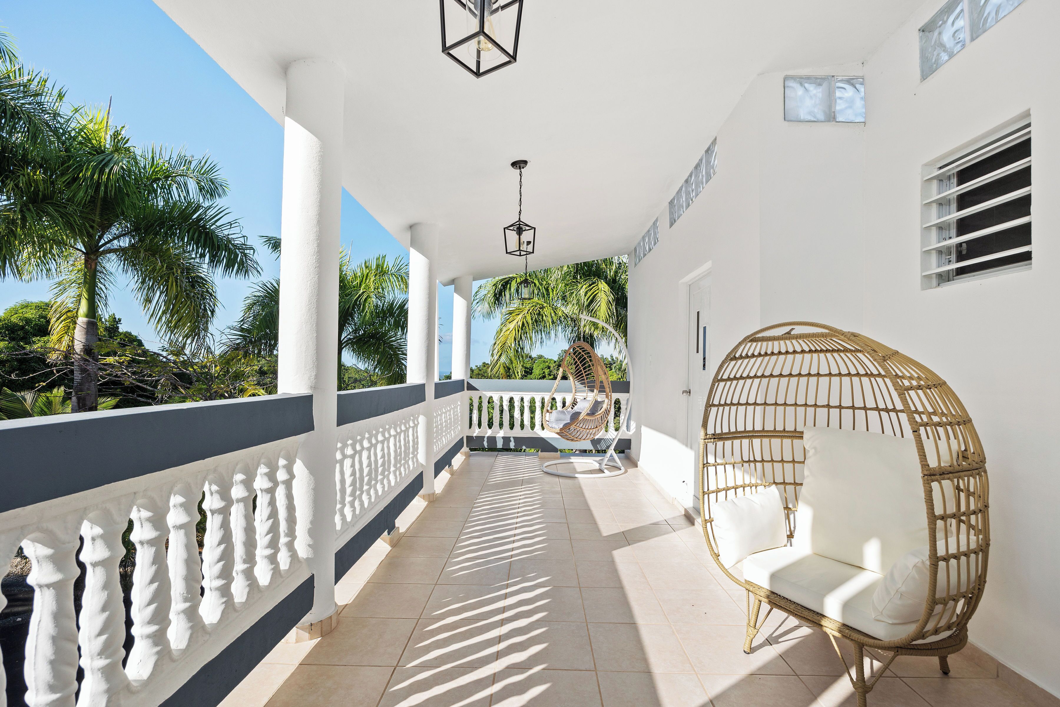 A covered balcony with a white railing, a wicker chair, and a view of palm trees.