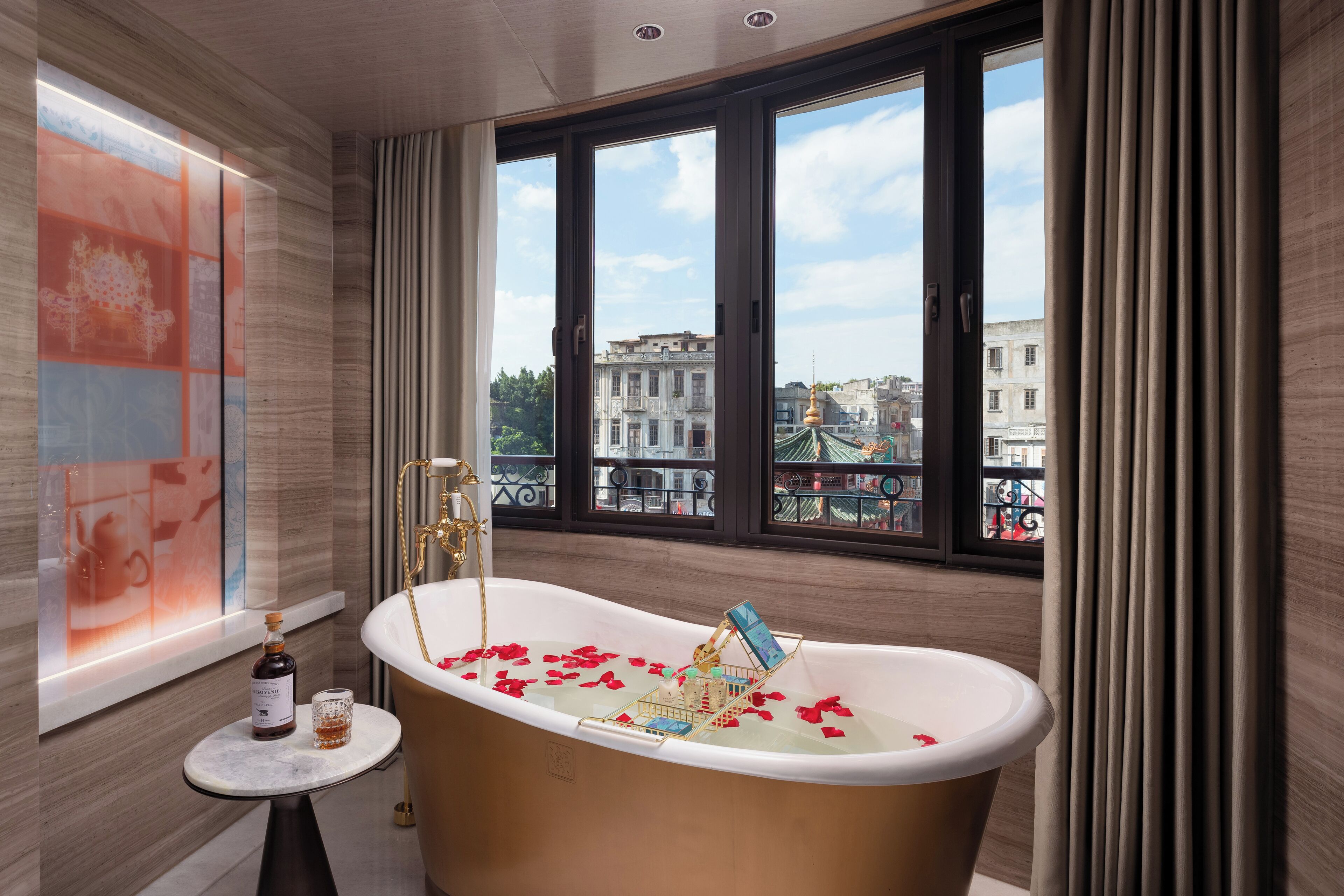 A bathroom with a freestanding bathtub, a small table with a bottle and glass, and a window with a city view.