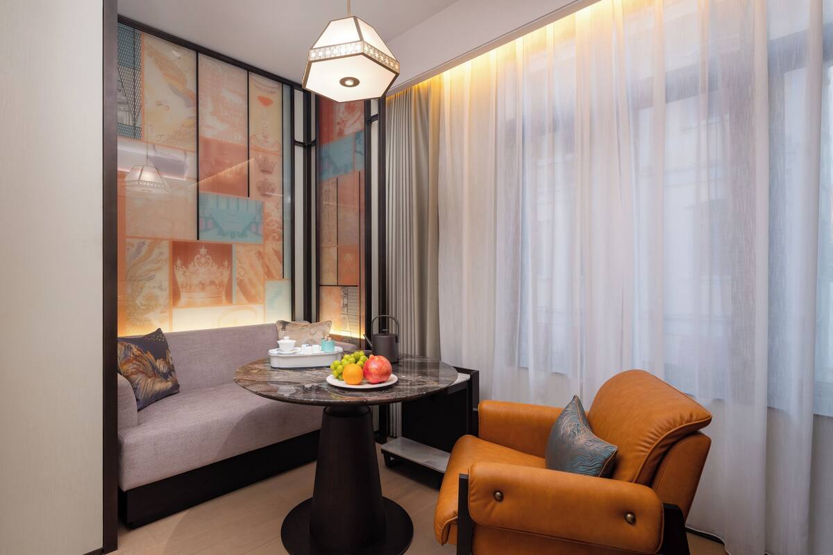 Habitación de hotel moderna con sofá, una mesa redonda con frutas, una cafetera y una lámpara.