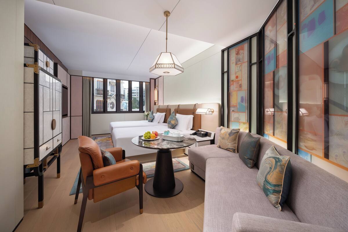 Habitación de hotel moderna con cama, sofá, mesa de comedor y una silla. Un ventanal grande ofrece vistas a la ciudad.
