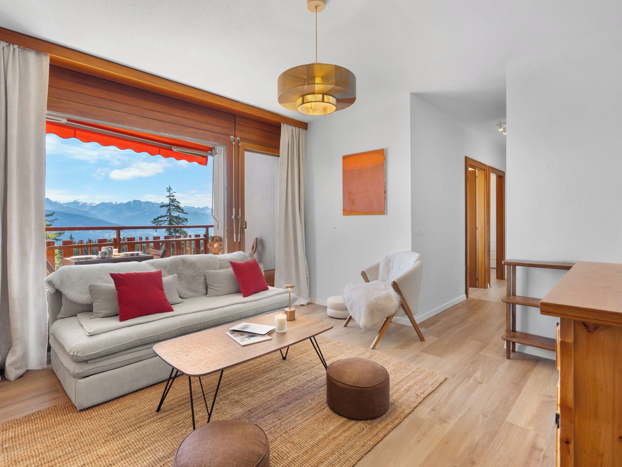 Uma sala de estar moderna com sofá, mesa de centro e poltrona, uma mesa de jantar de madeira e vista para montanhas através de uma varanda.