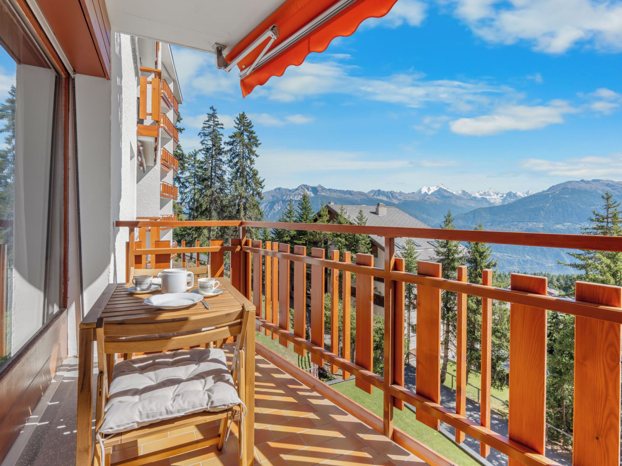 En balkon med et træbord og stole, udsigt til bjerge, og et gelænder af træ.
