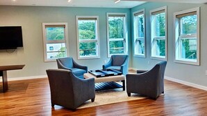 A spacious living room with a wooden floor, large windows, and a flat-screen TV mounted on the wall.