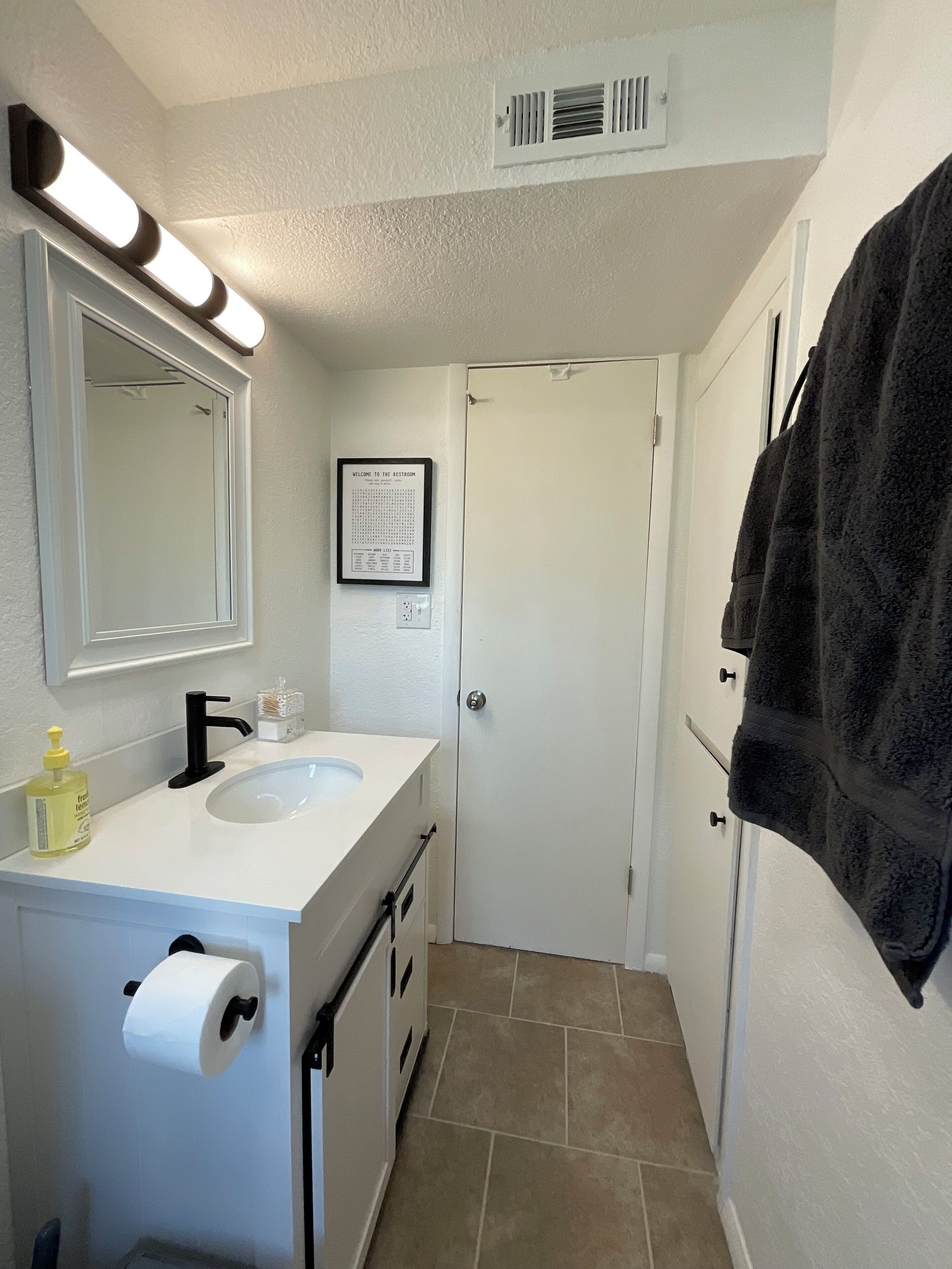 Une salle de bain avec un lavabo blanc, un miroir, un panneau d’accueil encadré, un porte-serviettes et un support à papier toilette.