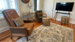 Een woonkamer met een bruine leren fauteuil, een groene comfortstoel, een houten salontafel, een grote klok, een televisietoestel en een kamerkleed met een patroon.