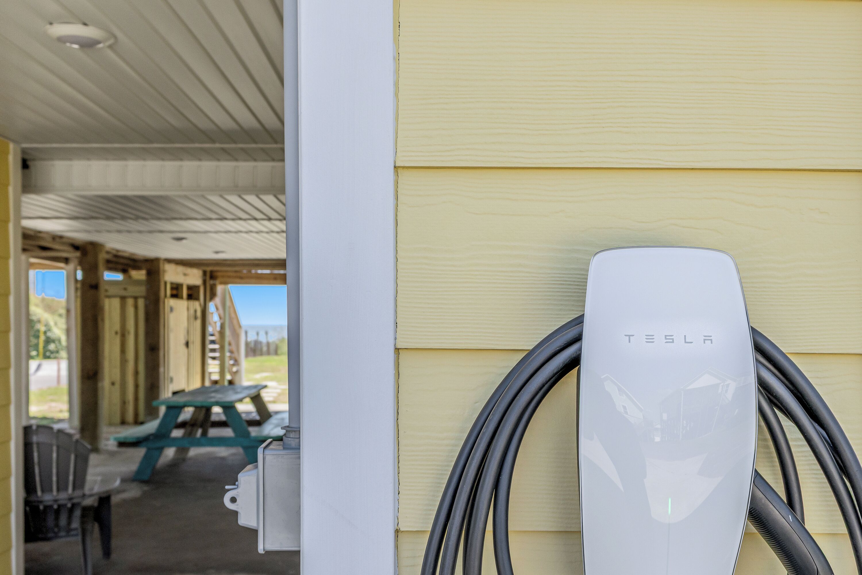 Une borne de recharge Tesla fixée à un bâtiment, avec un espace extérieur abrité comprenant une table de pique-nique et des chaises.