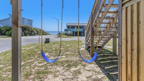 Eine Holzschaukel mit blauen Sitzen, eine hölzerne Konstruktion und im Hintergrund ein Gebäude sowie eine Straße.