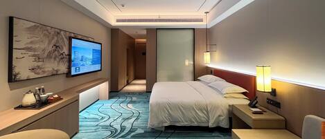 A hotel room with a bed, a desk, a chair, a TV, and a wall-mounted lamp.