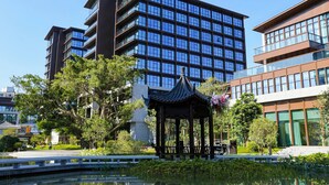 Un edificio moderno de varios pisos y grandes ventanales, rodeado de vegetación y un pabellón de estilo tradicional.