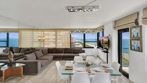 A modern living room with a large sectional sofa, a glass dining table set for four, and a view of the ocean. - Stylish Oceanfront 2 BR 1.5 BA Condo with Resort Amenities, Pools, Gym, Sauna 1402 (Miami Beach)